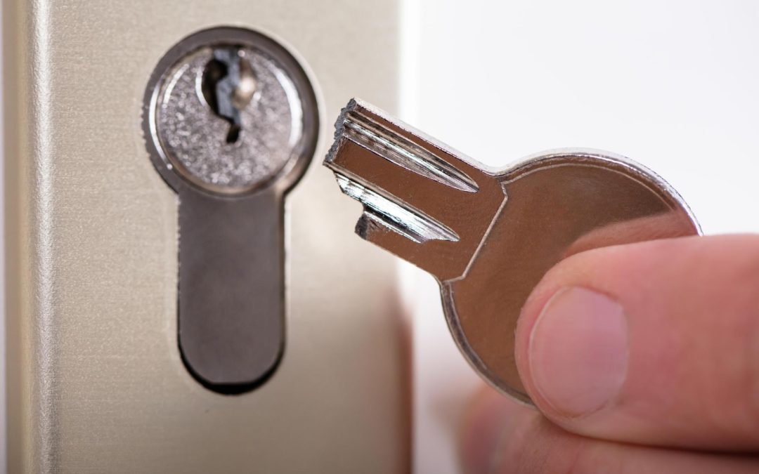 A hand holding a broken key near a door lock, with part of the key stuck inside the lock.